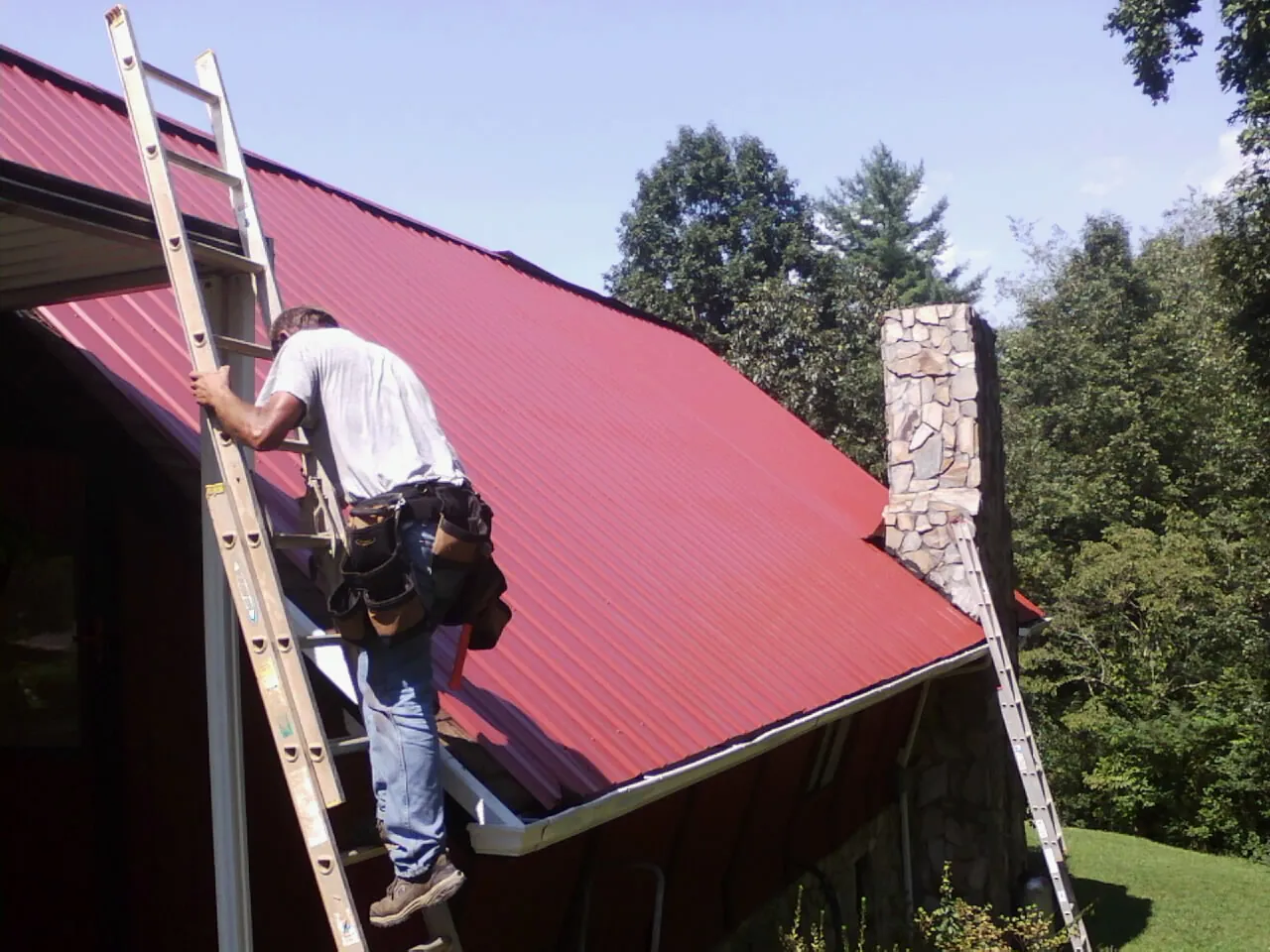 Roofer on a ladder beside a red metal roof and stone chimney — Soffit & Fascia Repair by Roofing Brothers in Callender